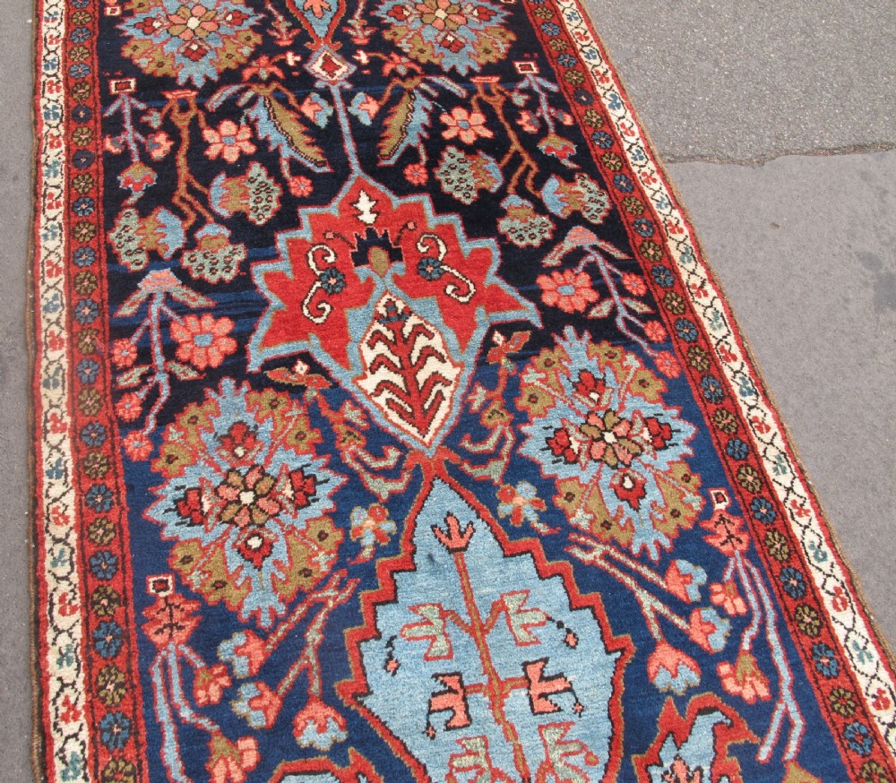 A long, narrow hand-knotted Persian wool runner featuring intricate geometric floral medallions in shades of navy blue, terracotta red, and sky blue along a dark central field. View XIV