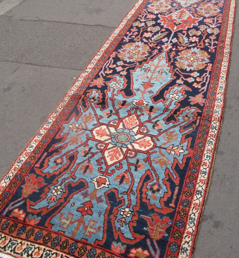 A long, narrow hand-knotted Persian wool runner featuring intricate geometric floral medallions in shades of navy blue, terracotta red, and sky blue along a dark central field. View XV