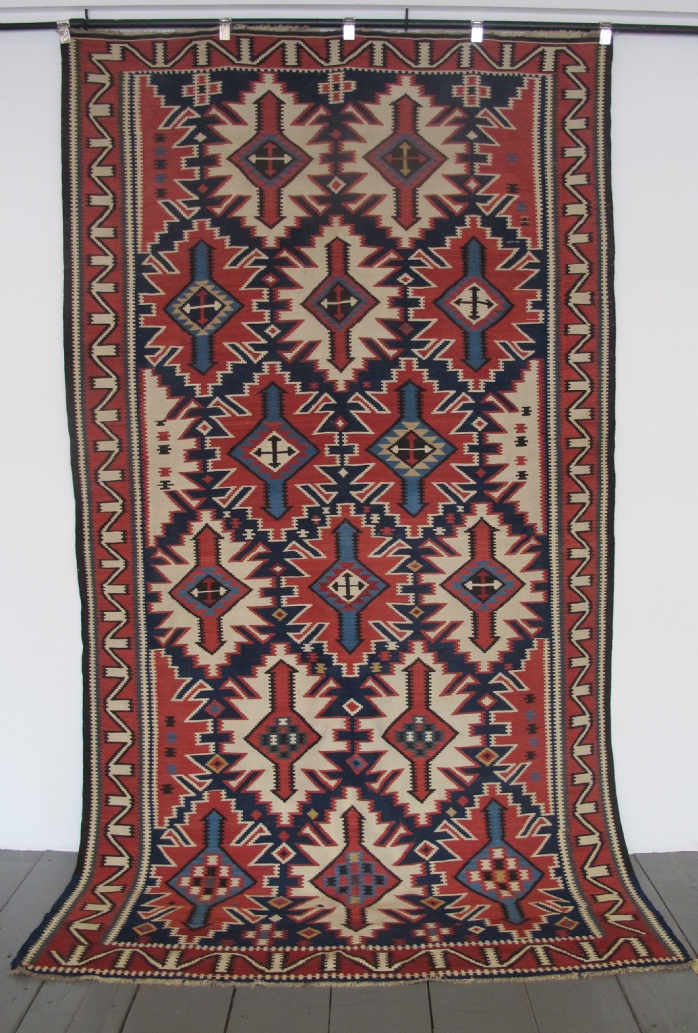 An antique Caucasian kilim flatweave wool rug displaying bold repeating diamond and cross motifs in terracotta red, navy blue, and cream.