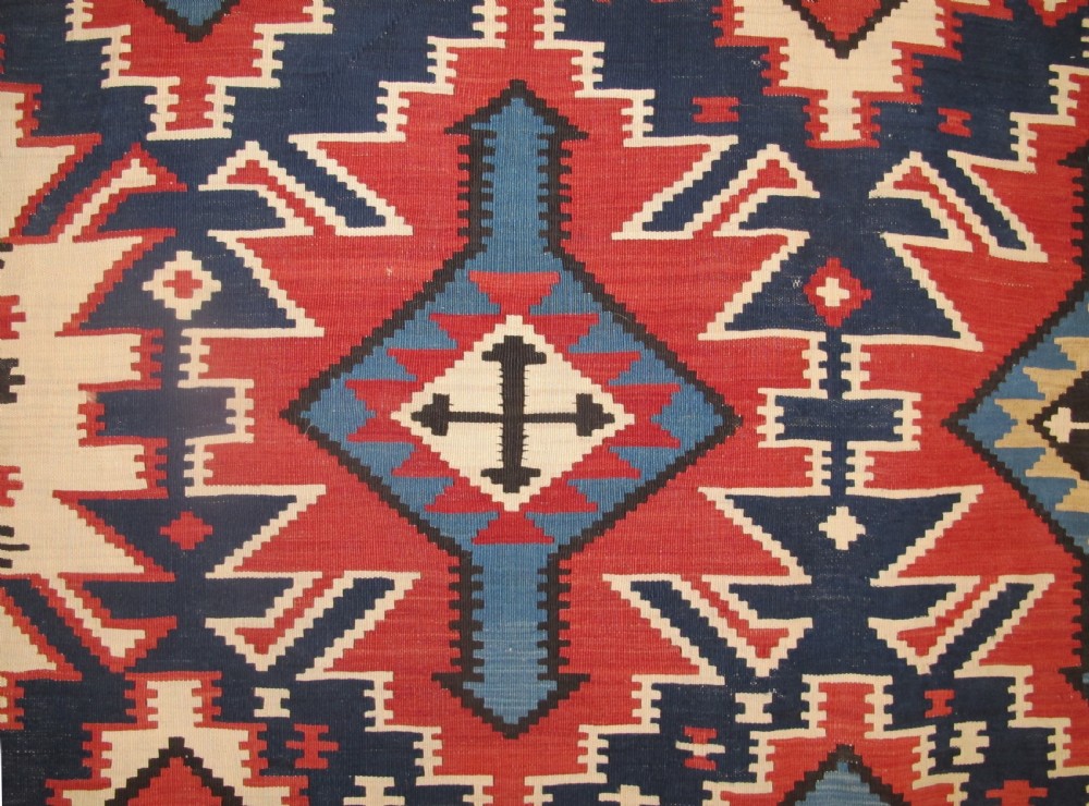 An antique Caucasian kilim flatweave wool rug displaying bold repeating diamond and cross motifs in terracotta red, navy blue, and cream. View III