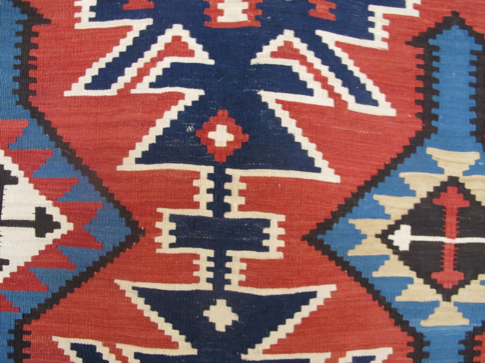 An antique Caucasian kilim flatweave wool rug displaying bold repeating diamond and cross motifs in terracotta red, navy blue, and cream. View IV