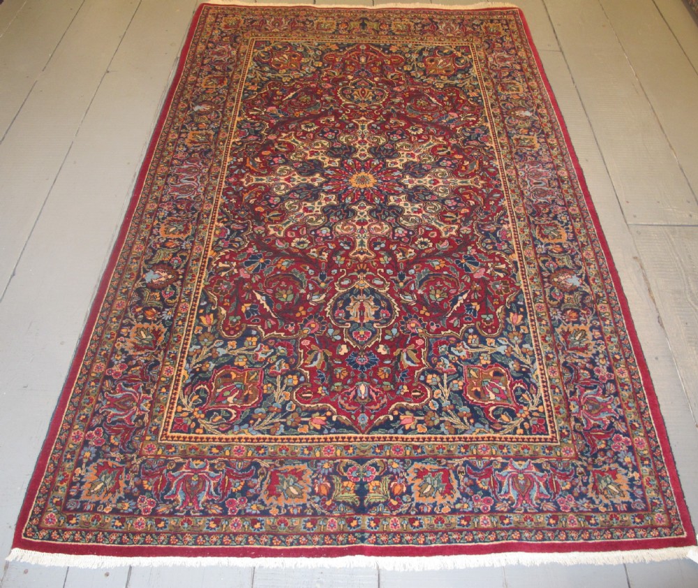 An antique Persian Meshed wool rug featuring an intricate central medallion surrounded by dense floral motifs in crimson red, navy blue, and cream.