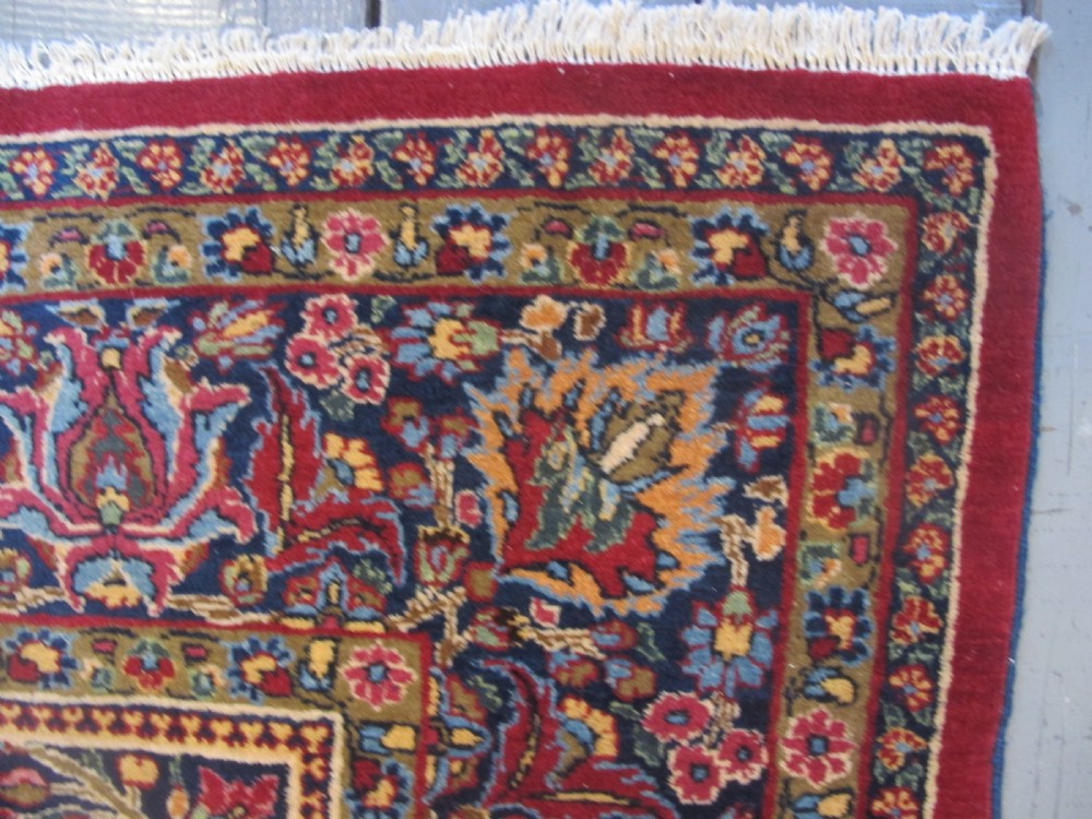 An antique Persian Meshed wool rug featuring an intricate central medallion surrounded by dense floral motifs in crimson red, navy blue, and cream. View II