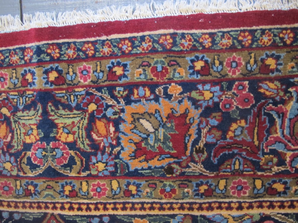 An antique Persian Meshed wool rug featuring an intricate central medallion surrounded by dense floral motifs in crimson red, navy blue, and cream. View III