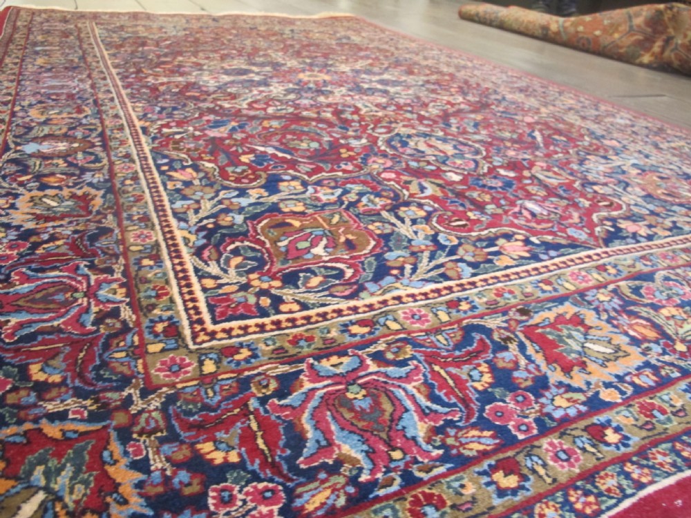 An antique Persian Meshed wool rug featuring an intricate central medallion surrounded by dense floral motifs in crimson red, navy blue, and cream. View IV