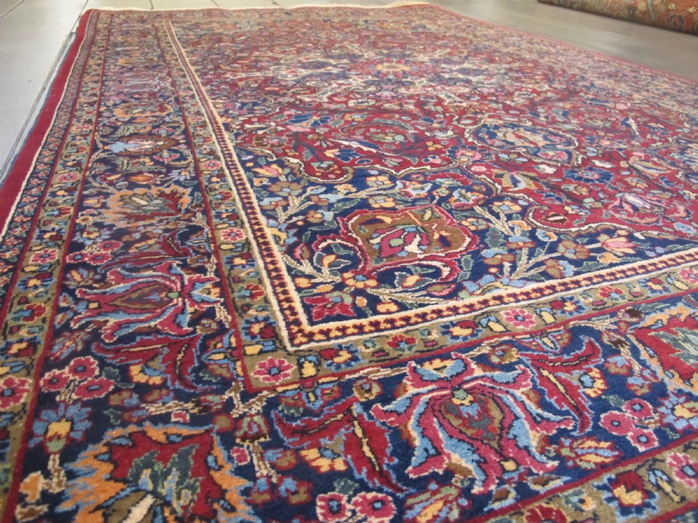 An antique Persian Meshed wool rug featuring an intricate central medallion surrounded by dense floral motifs in crimson red, navy blue, and cream. View V