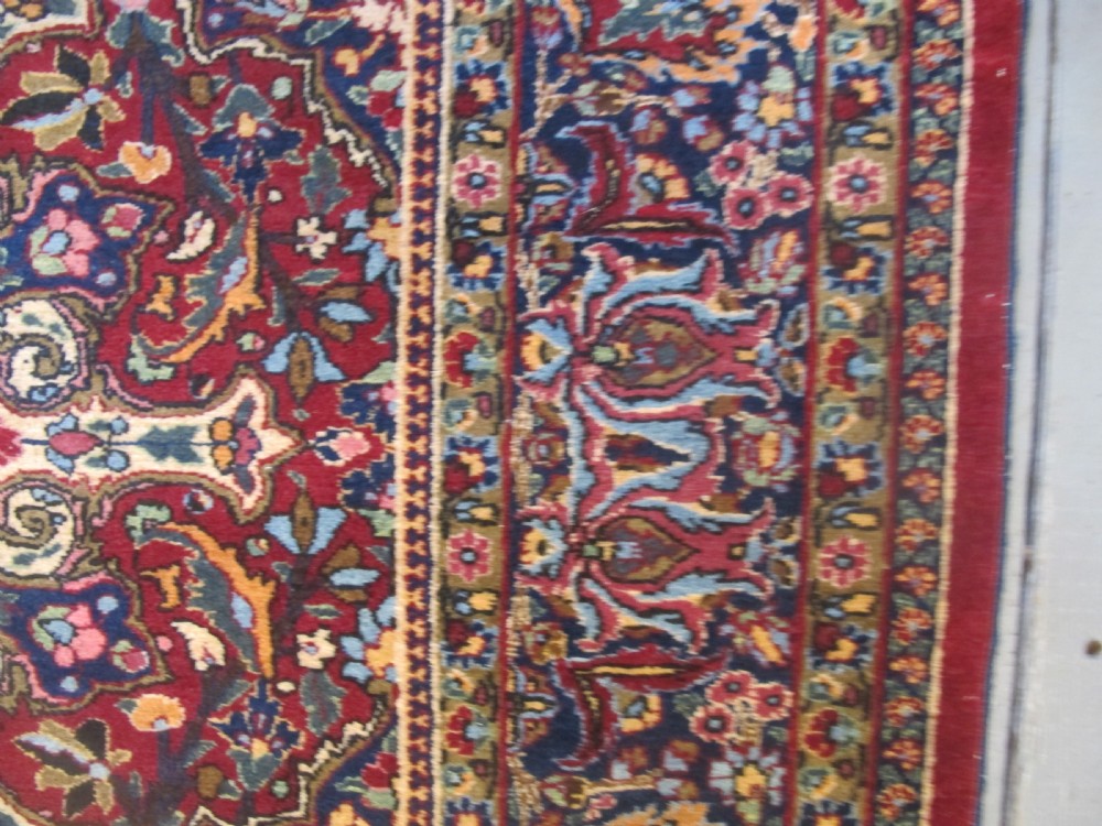 An antique Persian Meshed wool rug featuring an intricate central medallion surrounded by dense floral motifs in crimson red, navy blue, and cream. View VII