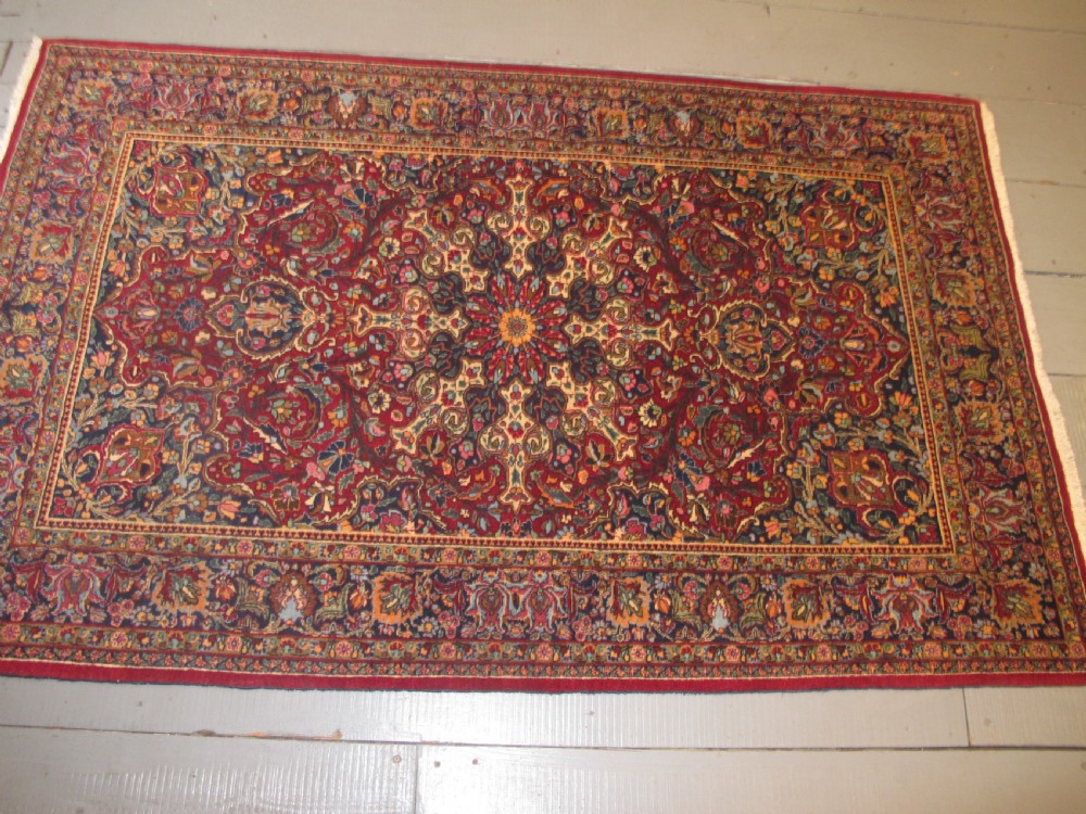 An antique Persian Meshed wool rug featuring an intricate central medallion surrounded by dense floral motifs in crimson red, navy blue, and cream. View IX