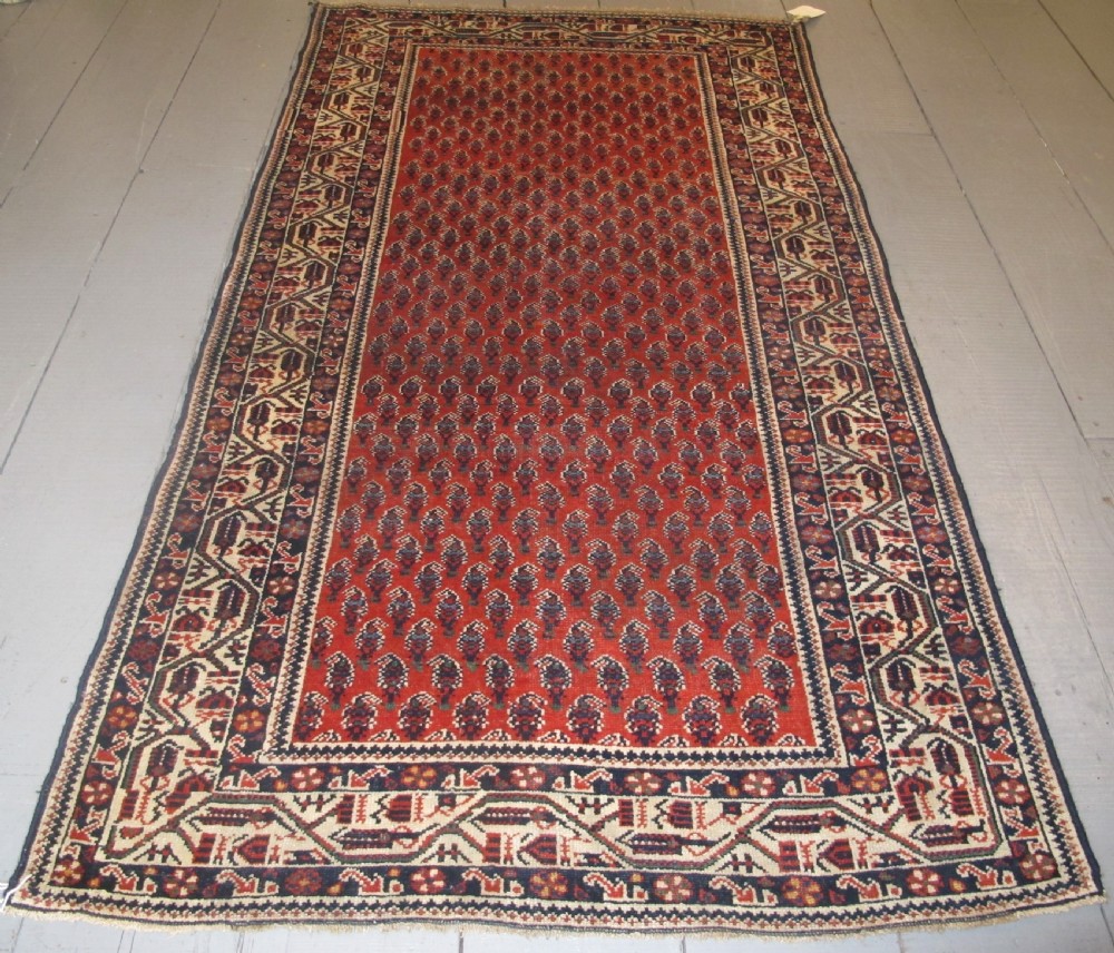 A rectangular hand-knotted wool rug featuring repeating boteh motifs on a red field enclosed by multi-layered cream and navy geometric borders.
