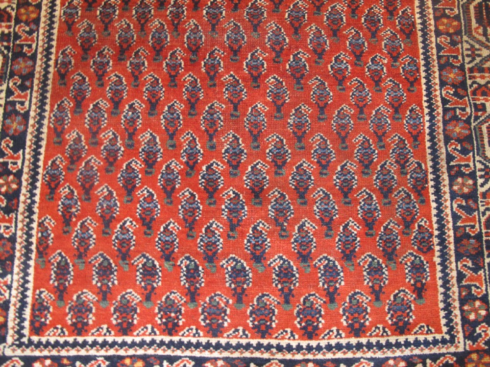 A rectangular hand-knotted wool rug featuring repeating boteh motifs on a red field enclosed by multi-layered cream and navy geometric borders. View IV