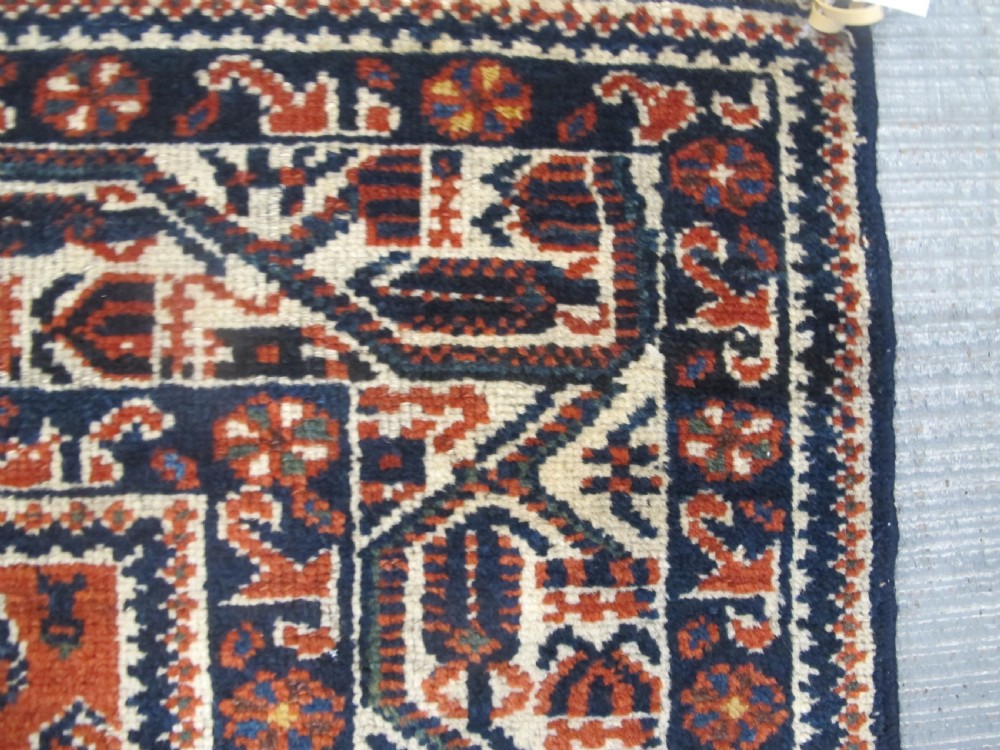 A rectangular hand-knotted wool rug featuring repeating boteh motifs on a red field enclosed by multi-layered cream and navy geometric borders. View V