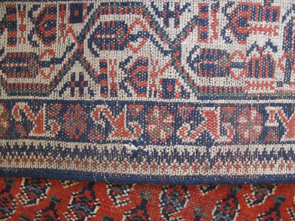 A rectangular hand-knotted wool rug featuring repeating boteh motifs on a red field enclosed by multi-layered cream and navy geometric borders. View VI