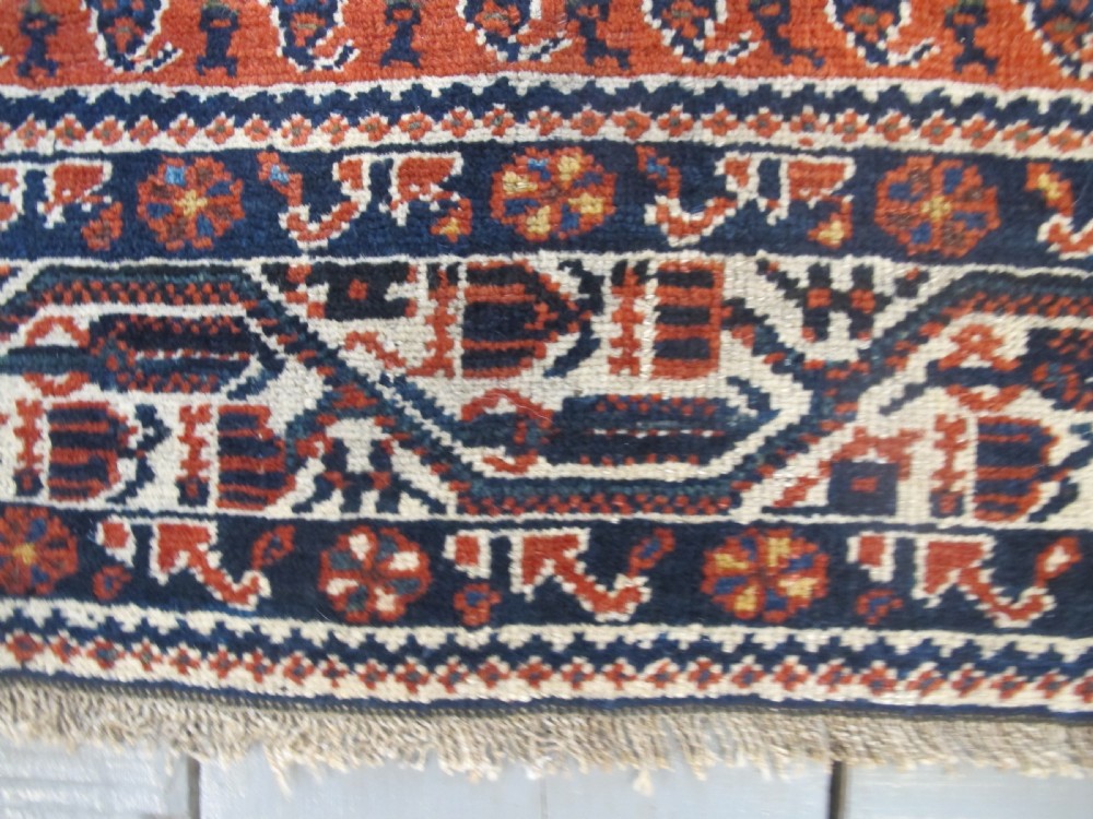A rectangular hand-knotted wool rug featuring repeating boteh motifs on a red field enclosed by multi-layered cream and navy geometric borders. View VII
