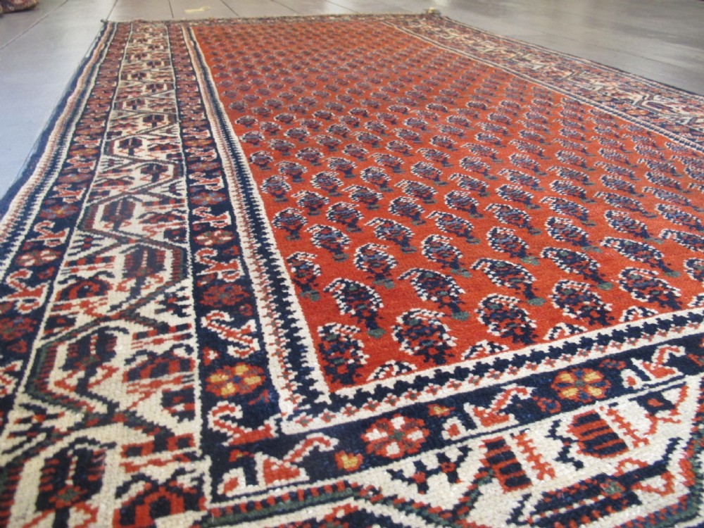 A rectangular hand-knotted wool rug featuring repeating boteh motifs on a red field enclosed by multi-layered cream and navy geometric borders. View IX