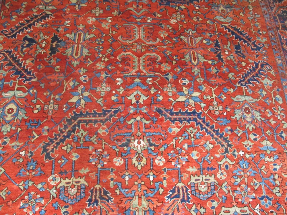 A large antique hand-knotted Persian Karaja wool carpet featuring a dominant red field with complex geometric floral medallions and a wide navy blue border. View IV