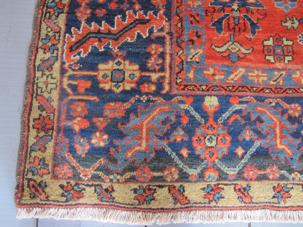 A large antique hand-knotted Persian Karaja wool carpet featuring a dominant red field with complex geometric floral medallions and a wide navy blue border. View V