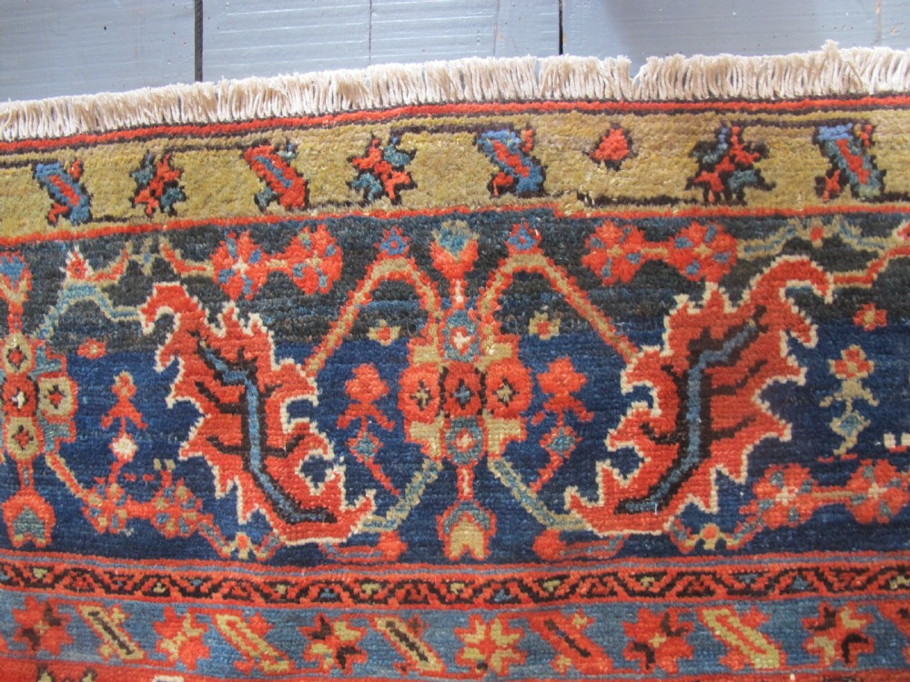 A large antique hand-knotted Persian Karaja wool carpet featuring a dominant red field with complex geometric floral medallions and a wide navy blue border. View VI