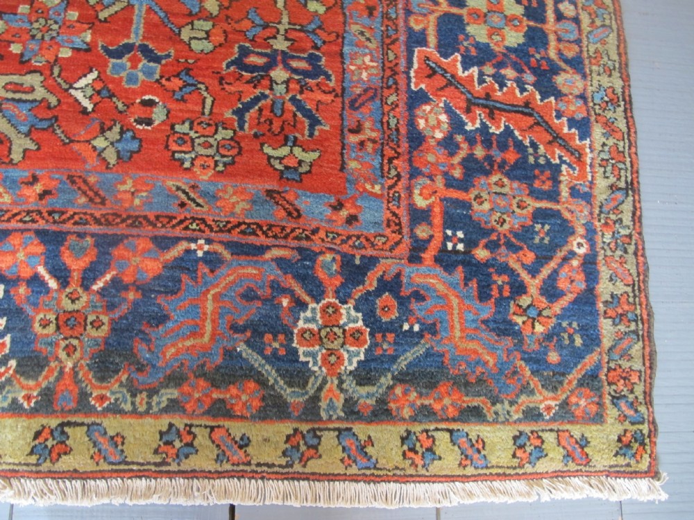 A large antique hand-knotted Persian Karaja wool carpet featuring a dominant red field with complex geometric floral medallions and a wide navy blue border. View VII