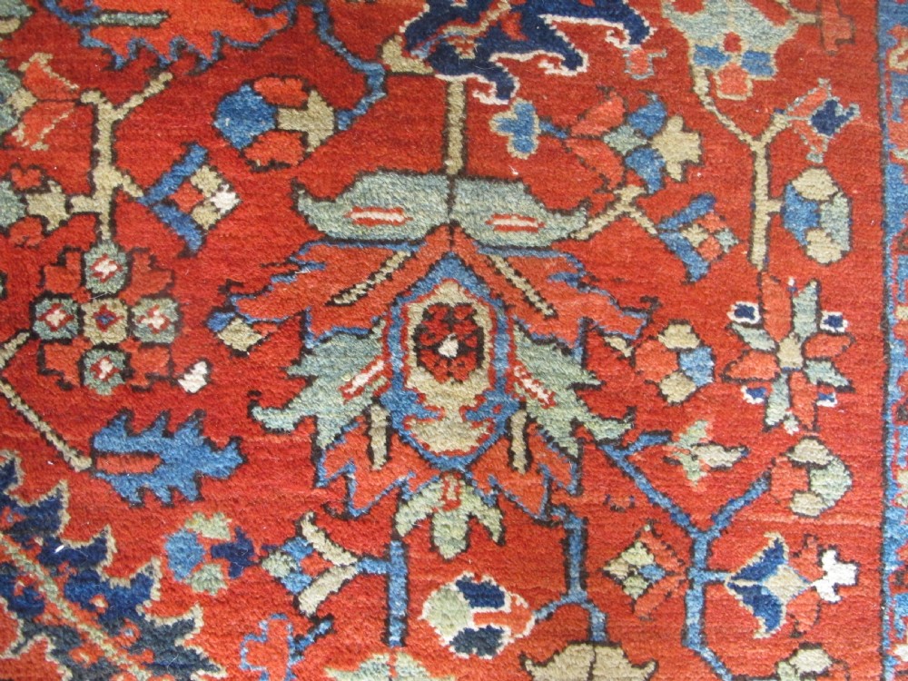 A large antique hand-knotted Persian Karaja wool carpet featuring a dominant red field with complex geometric floral medallions and a wide navy blue border. View IX