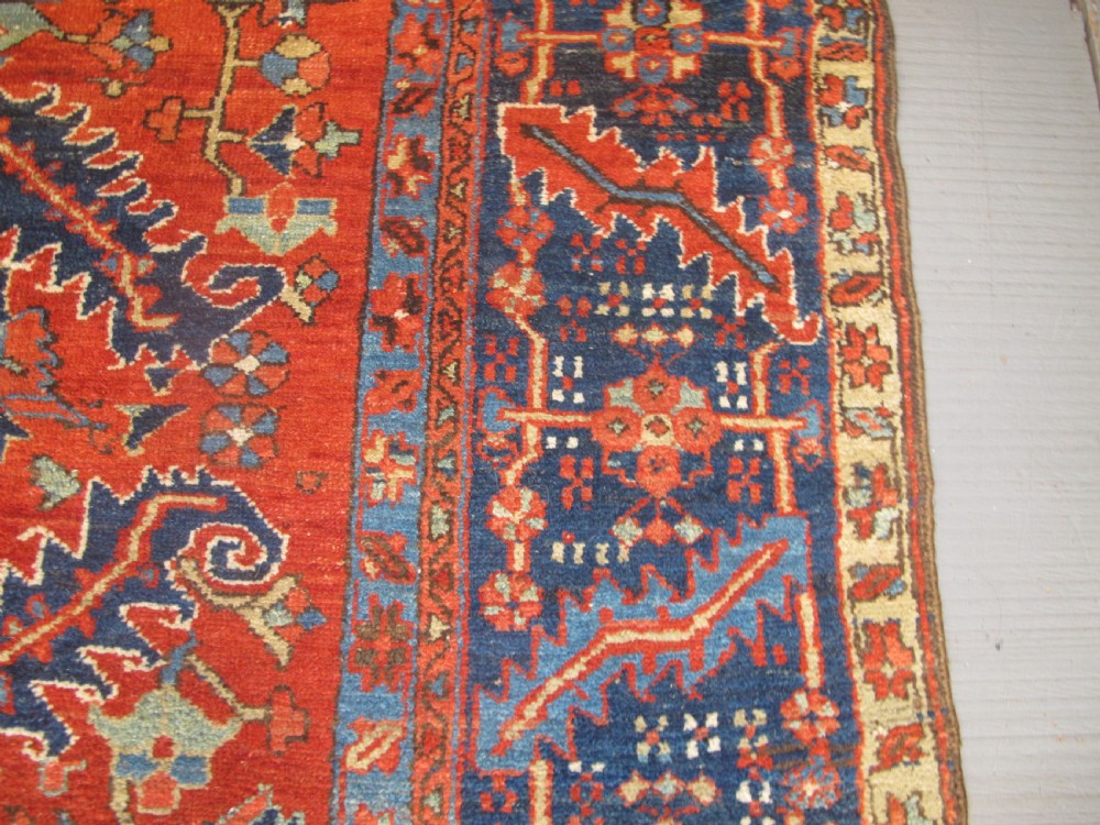 A large antique hand-knotted Persian Karaja wool carpet featuring a dominant red field with complex geometric floral medallions and a wide navy blue border. View X