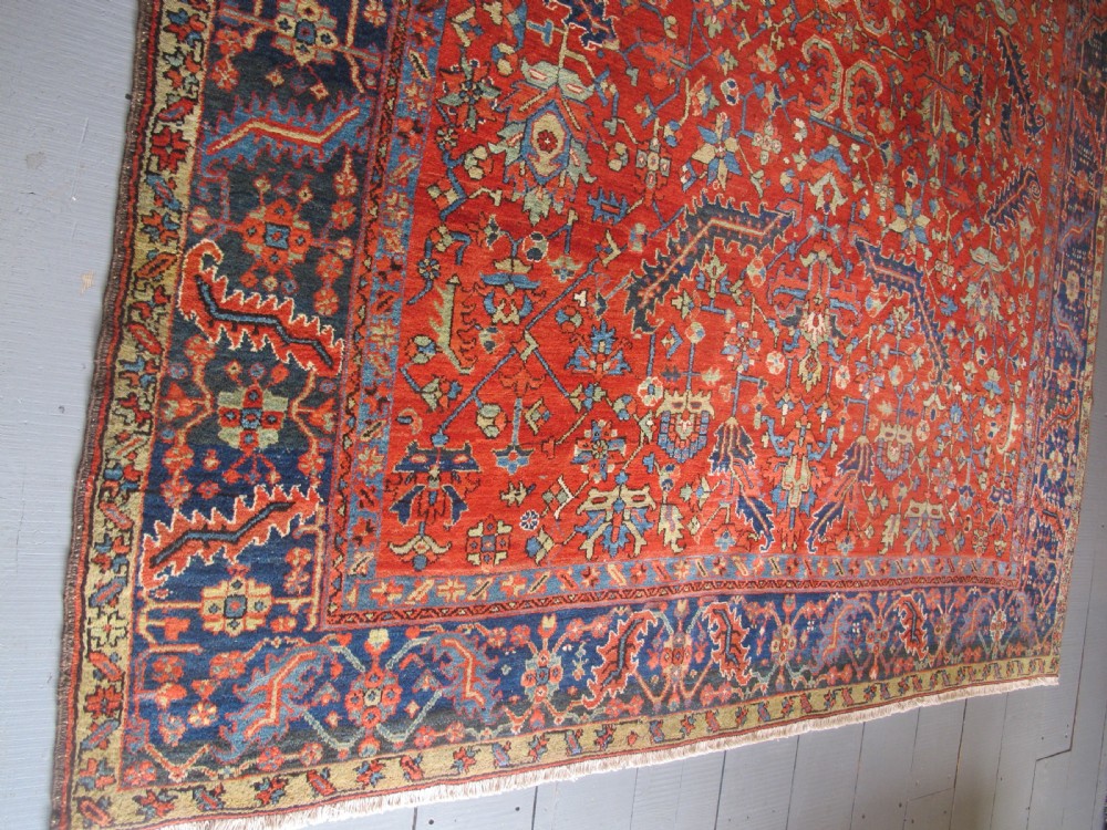 A large antique hand-knotted Persian Karaja wool carpet featuring a dominant red field with complex geometric floral medallions and a wide navy blue border. View XIV