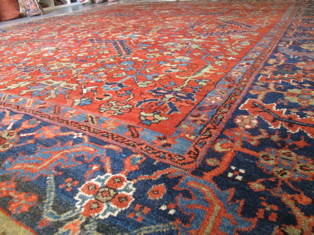 A large antique hand-knotted Persian Karaja wool carpet featuring a dominant red field with complex geometric floral medallions and a wide navy blue border. View XV