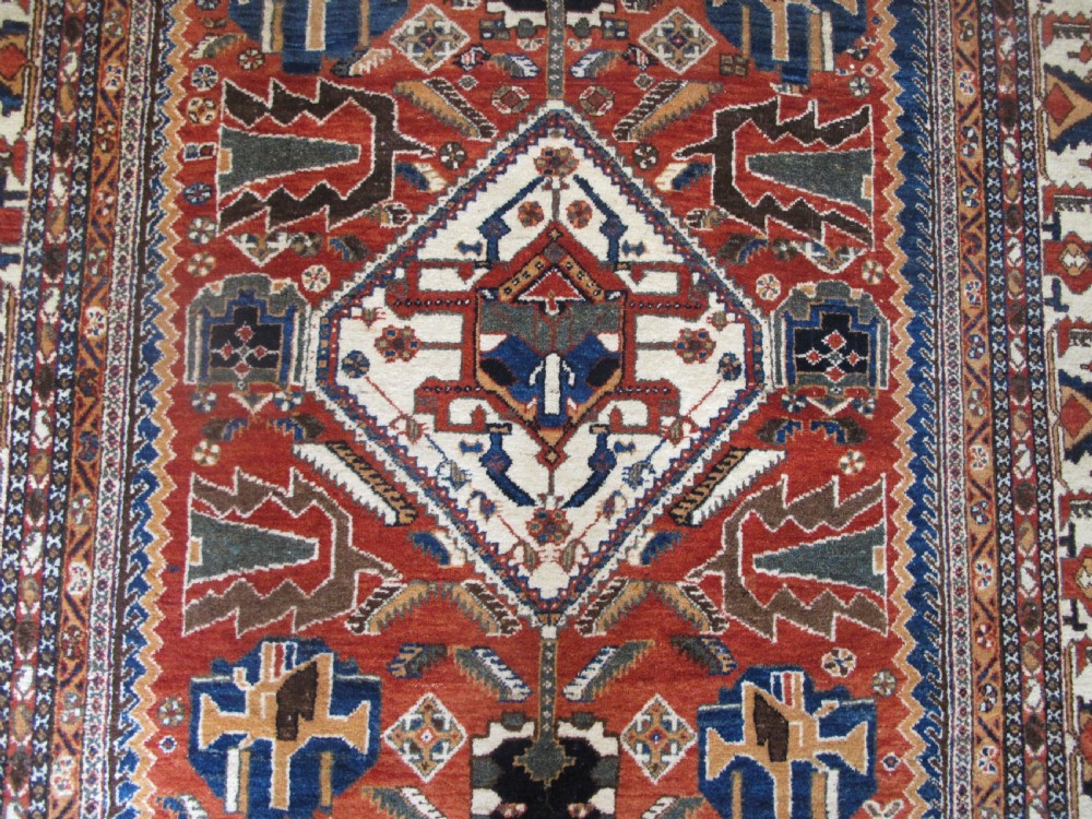 An antique South Persian hand-knotted wool rug featuring a central ivory diamond medallion within a terracotta field, bordered by indigo corners and intricate geometric bands. View IX