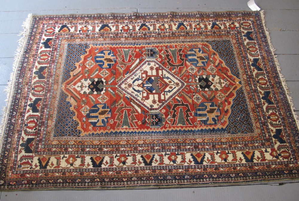 An antique South Persian hand-knotted wool rug featuring a central ivory diamond medallion within a terracotta field, bordered by indigo corners and intricate geometric bands. View XIII
