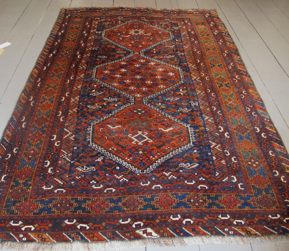 An antique hand-knotted Persian tribal rug featuring three central diamond medallions surrounded by geometric patterns in deep red, navy blue, and terracotta tones.