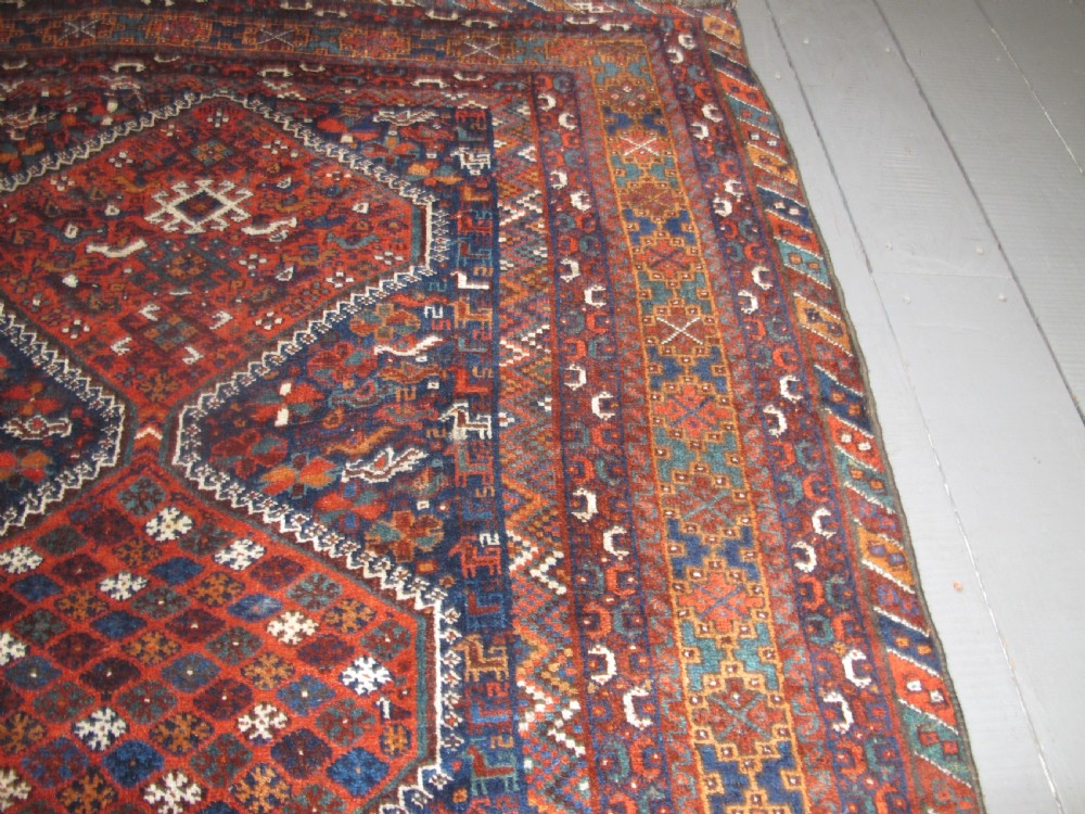 An antique hand-knotted Persian tribal rug featuring three central diamond medallions surrounded by geometric patterns in deep red, navy blue, and terracotta tones. View X