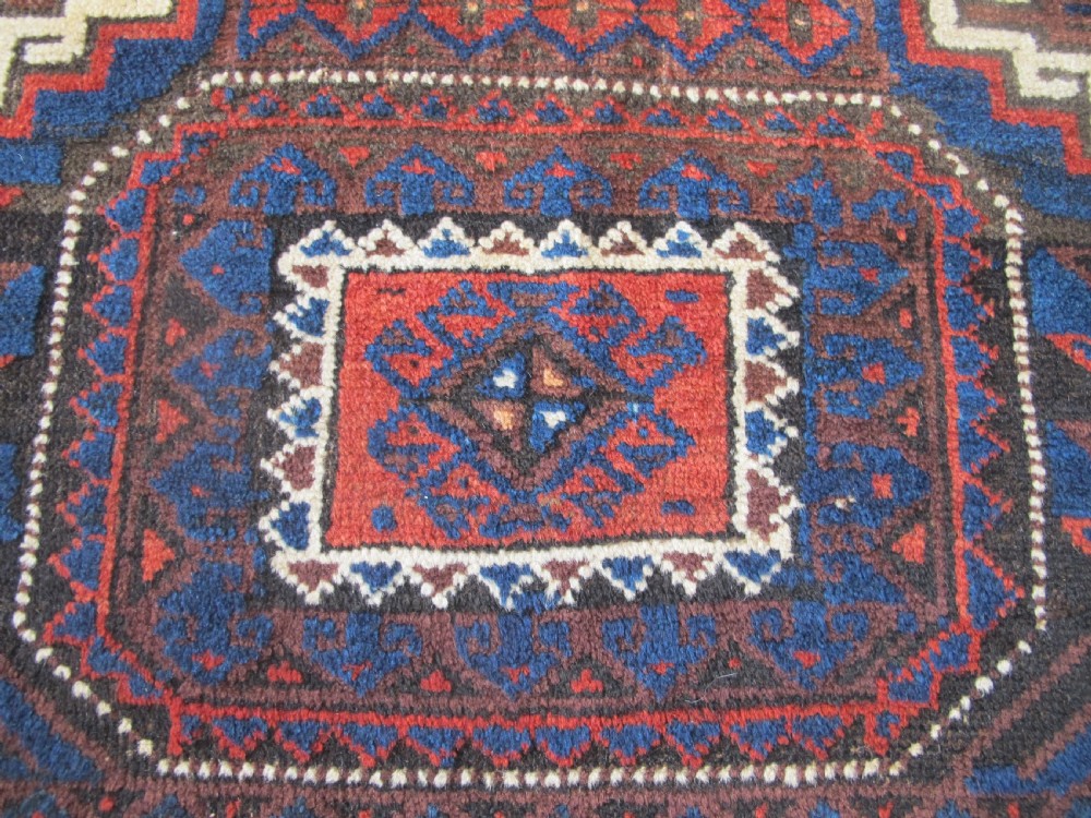 An antique hand-woven Belouch wool rug featuring geometric octagonal medallions in deep red, navy blue, and dark brown tones. View V