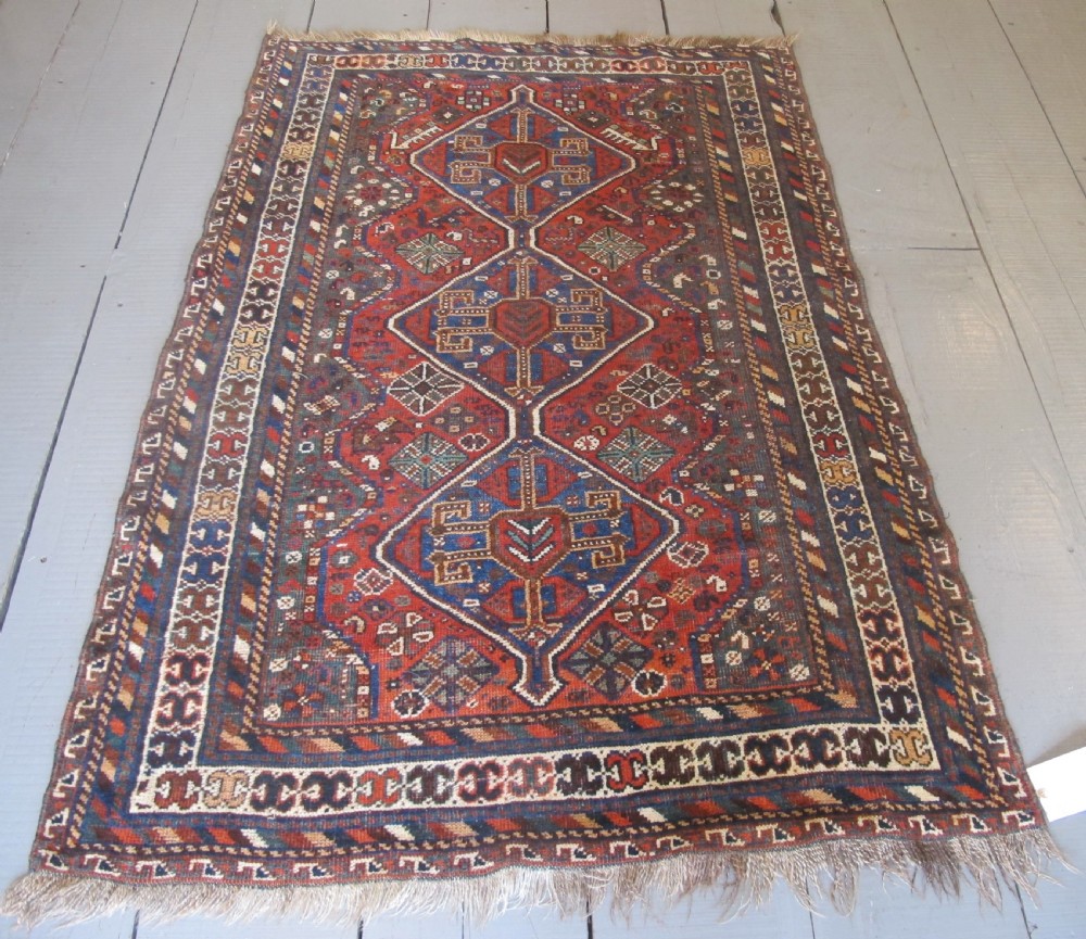 A hand-knotted antique Persian Shiraz wool rug featuring three geometric diamond medallions in blue and cream on a deep terracotta red field with decorative tribal borders. View III