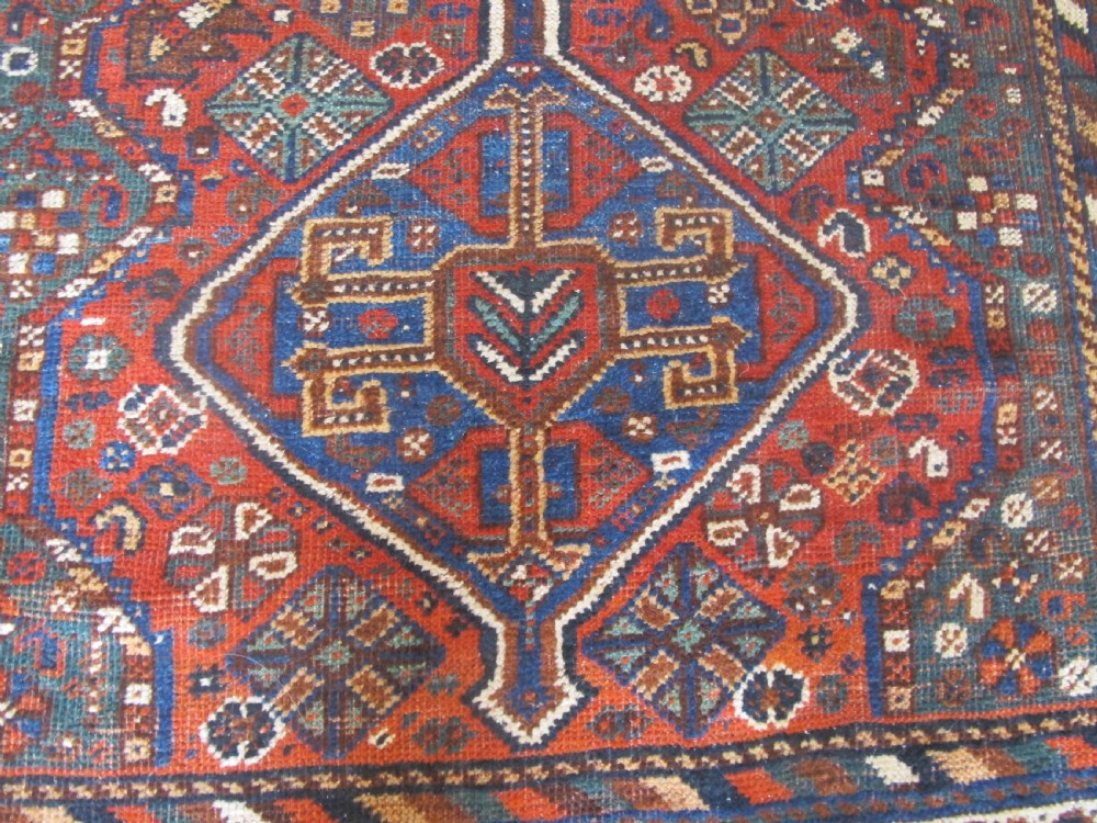 A hand-knotted antique Persian Shiraz wool rug featuring three geometric diamond medallions in blue and cream on a deep terracotta red field with decorative tribal borders. View VI
