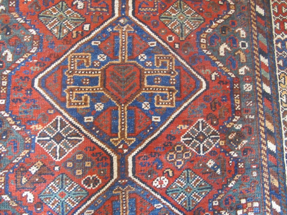 A hand-knotted antique Persian Shiraz wool rug featuring three geometric diamond medallions in blue and cream on a deep terracotta red field with decorative tribal borders. View VII