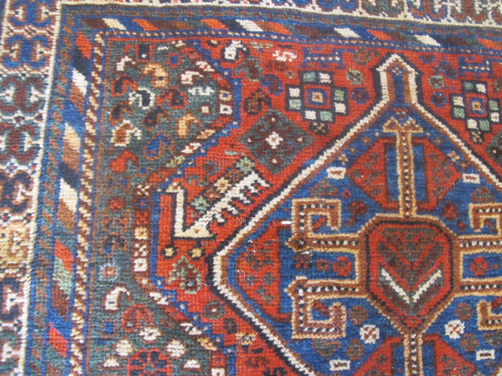 A hand-knotted antique Persian Shiraz wool rug featuring three geometric diamond medallions in blue and cream on a deep terracotta red field with decorative tribal borders. View IX