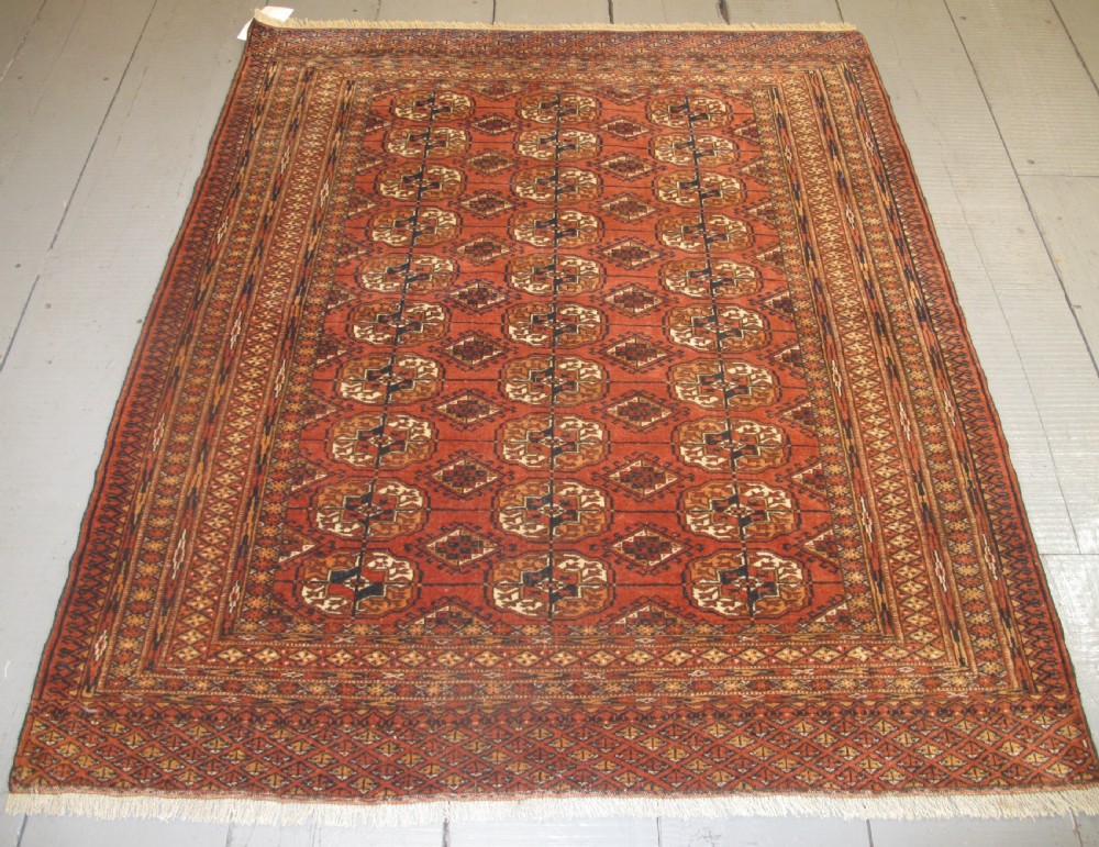 A rectangular hand-knotted Tekke rug featuring repeating octagonal gul motifs in dark blue and ivory across a terracotta red wool ground.