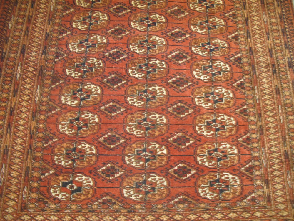 A rectangular hand-knotted Tekke rug featuring repeating octagonal gul motifs in dark blue and ivory across a terracotta red wool ground. View III