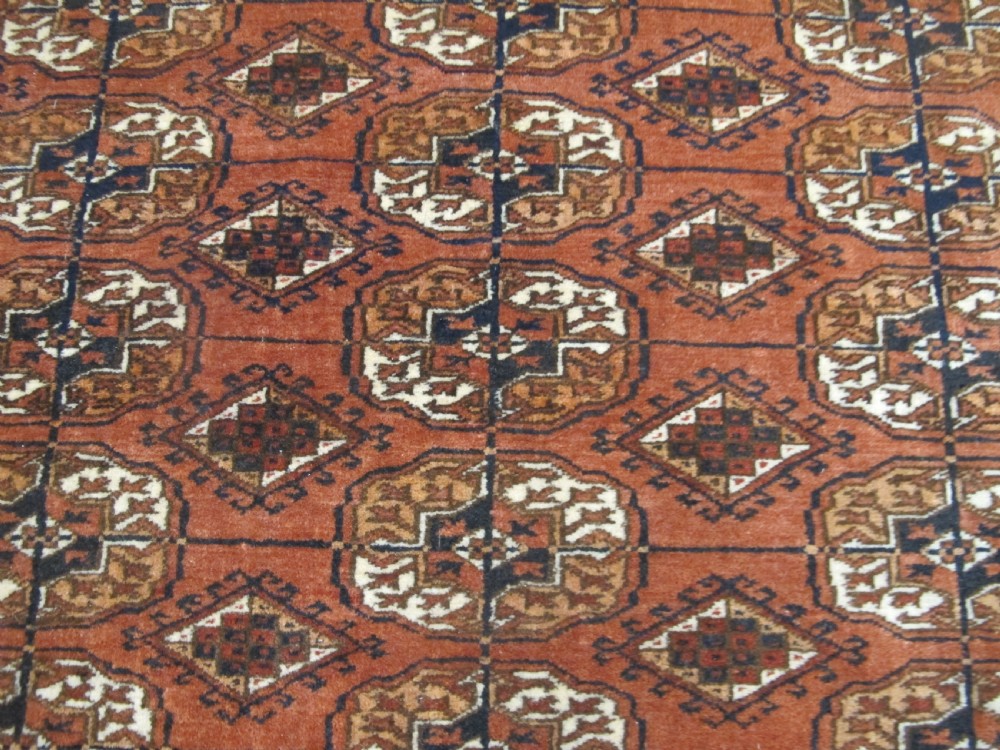A rectangular hand-knotted Tekke rug featuring repeating octagonal gul motifs in dark blue and ivory across a terracotta red wool ground. View IV