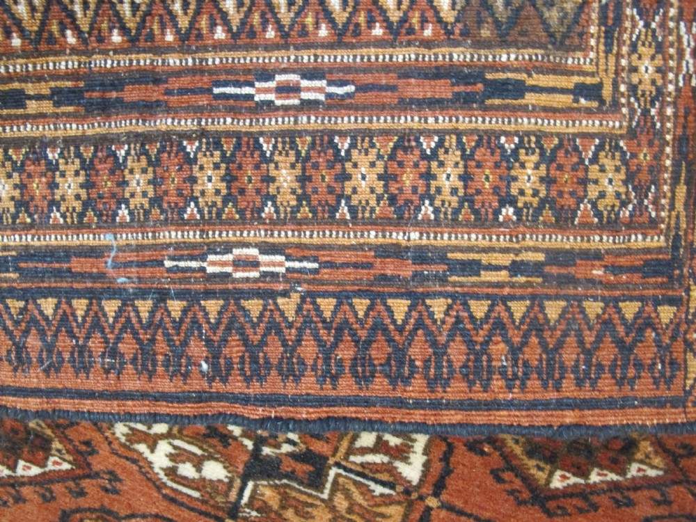 A rectangular hand-knotted Tekke rug featuring repeating octagonal gul motifs in dark blue and ivory across a terracotta red wool ground. View V