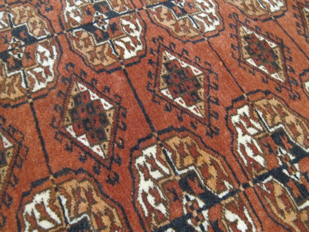 A rectangular hand-knotted Tekke rug featuring repeating octagonal gul motifs in dark blue and ivory across a terracotta red wool ground. View VII