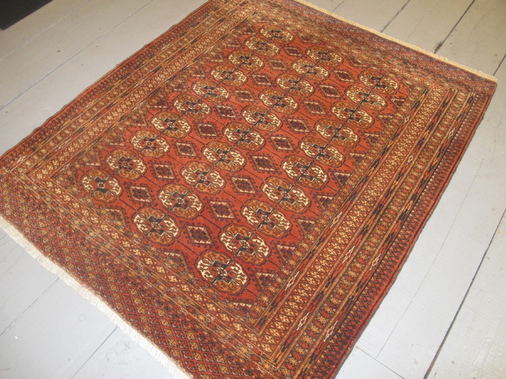 A rectangular hand-knotted Tekke rug featuring repeating octagonal gul motifs in dark blue and ivory across a terracotta red wool ground. View X