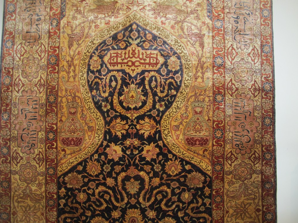 A mid-20th-century Turkish silk rug featuring a dark navy mihrab arch filled with floral motifs, surrounded by an ochre border with calligraphic script and bird illustrations. View VI