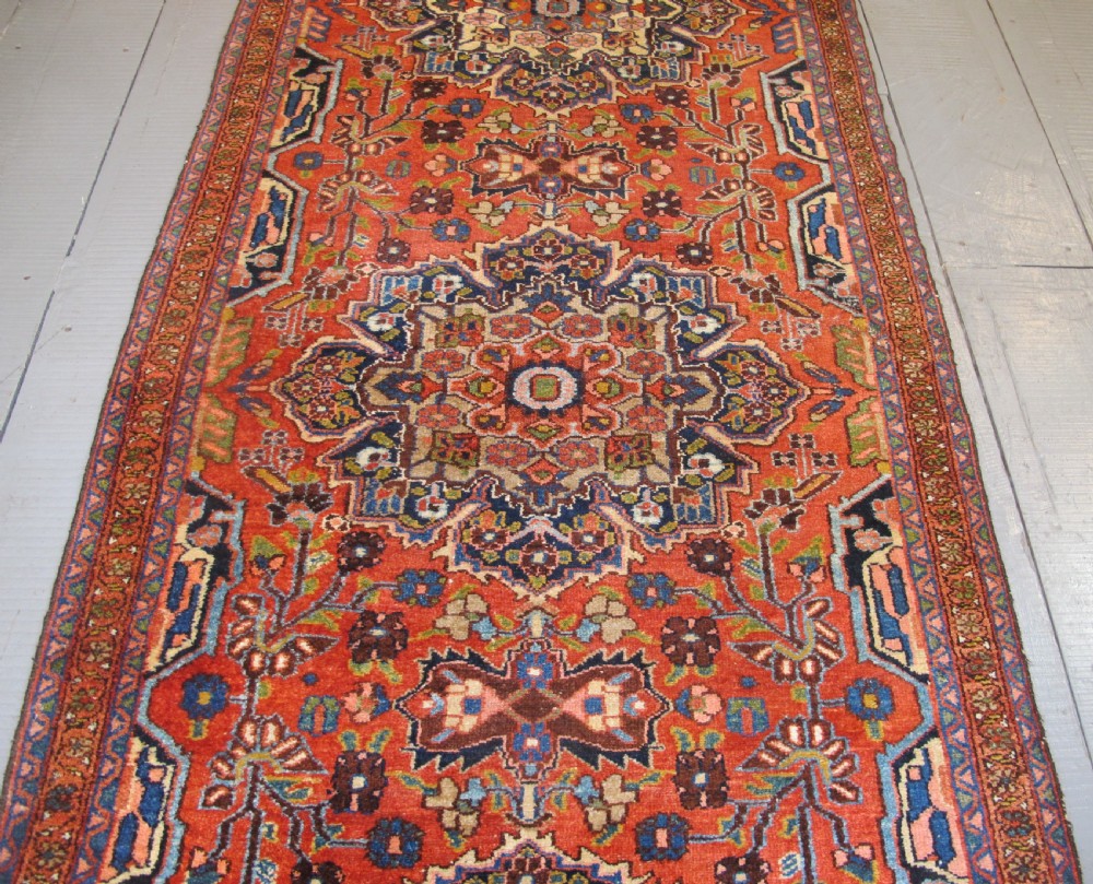 A long hand-knotted antique Persian Hamadan runner rug featuring terracotta red, navy blue, and cream floral medallion motifs throughout the central field. View III