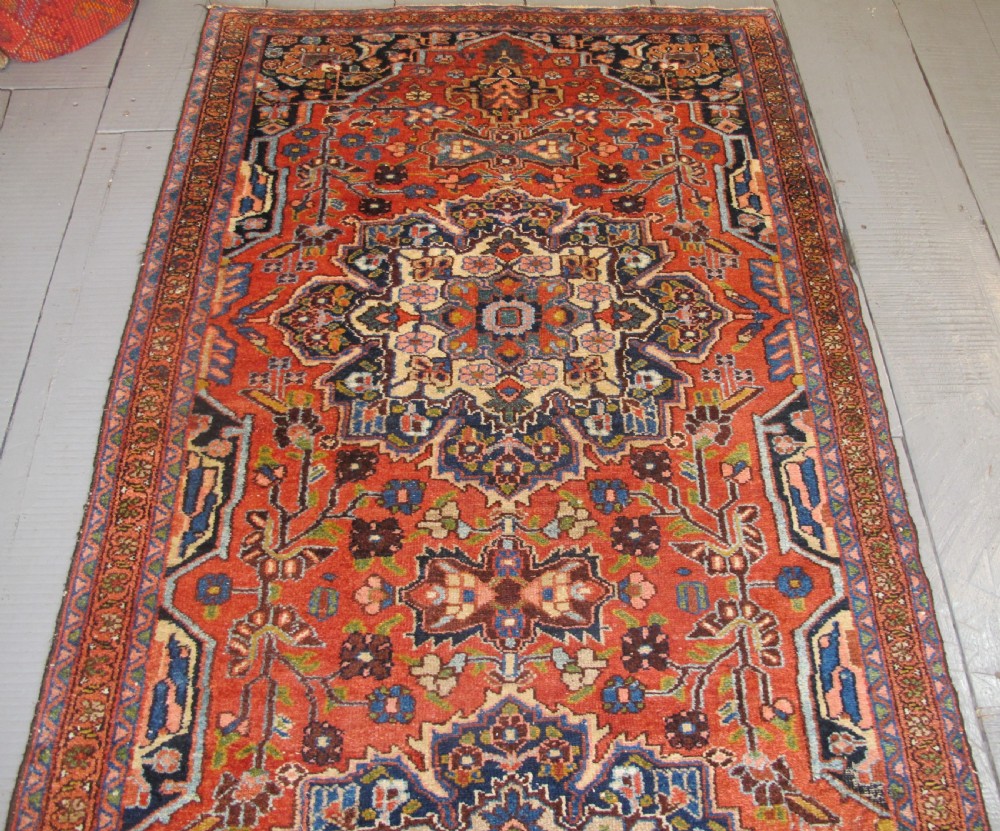 A long hand-knotted antique Persian Hamadan runner rug featuring terracotta red, navy blue, and cream floral medallion motifs throughout the central field. View IV