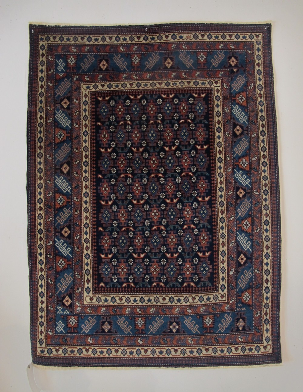 A rectangular hand-knotted wool rug featuring a dark navy central field filled with repeating geometric motifs surrounded by intricate multi-layered pattern borders in blue, terracotta, and cream.