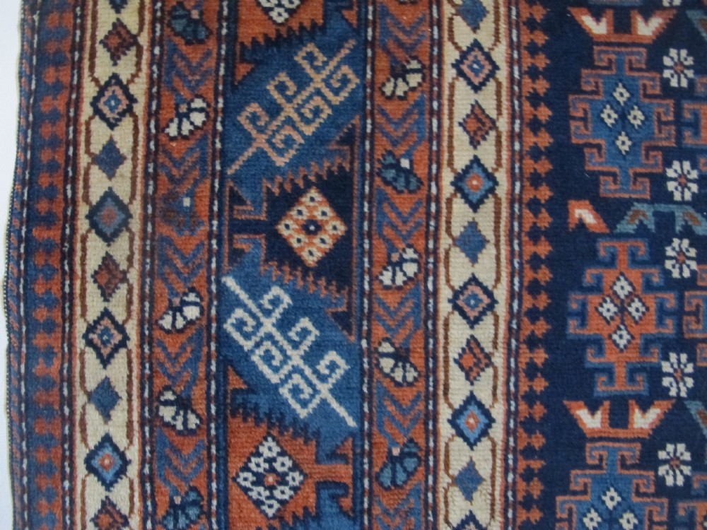A rectangular hand-knotted wool rug featuring a dark navy central field filled with repeating geometric motifs surrounded by intricate multi-layered pattern borders in blue, terracotta, and cream. View III