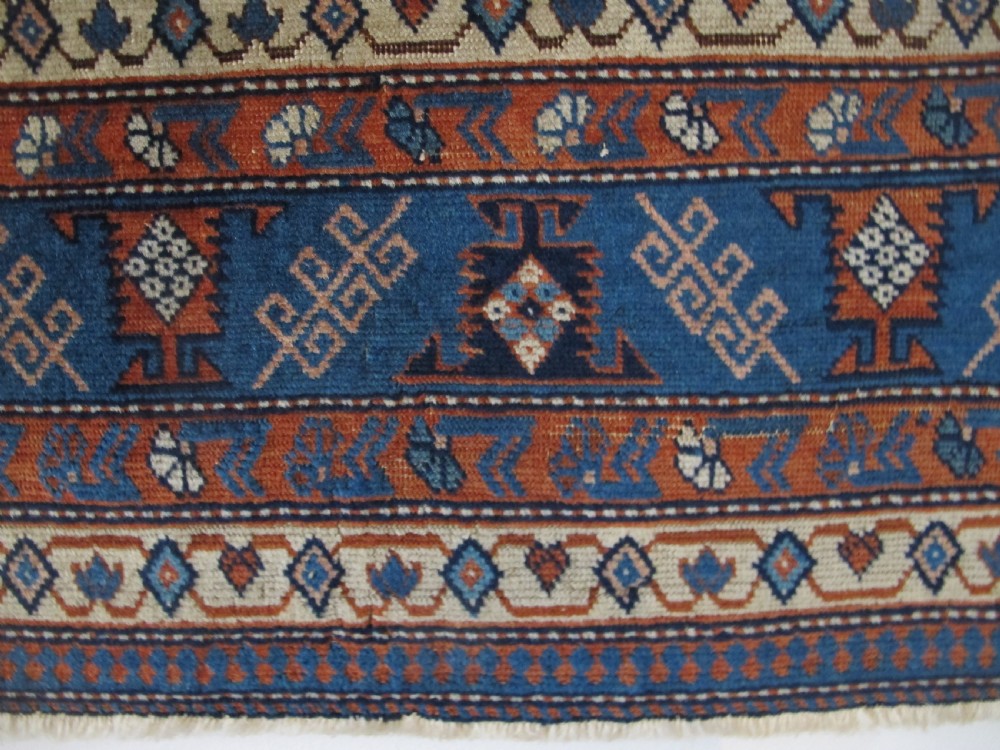 A rectangular hand-knotted wool rug featuring a dark navy central field filled with repeating geometric motifs surrounded by intricate multi-layered pattern borders in blue, terracotta, and cream. View V