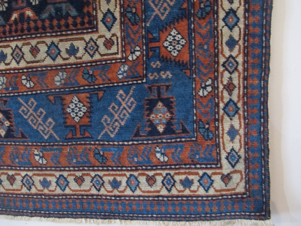 A rectangular hand-knotted wool rug featuring a dark navy central field filled with repeating geometric motifs surrounded by intricate multi-layered pattern borders in blue, terracotta, and cream. View VI