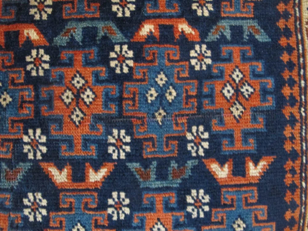 A rectangular hand-knotted wool rug featuring a dark navy central field filled with repeating geometric motifs surrounded by intricate multi-layered pattern borders in blue, terracotta, and cream. View VII
