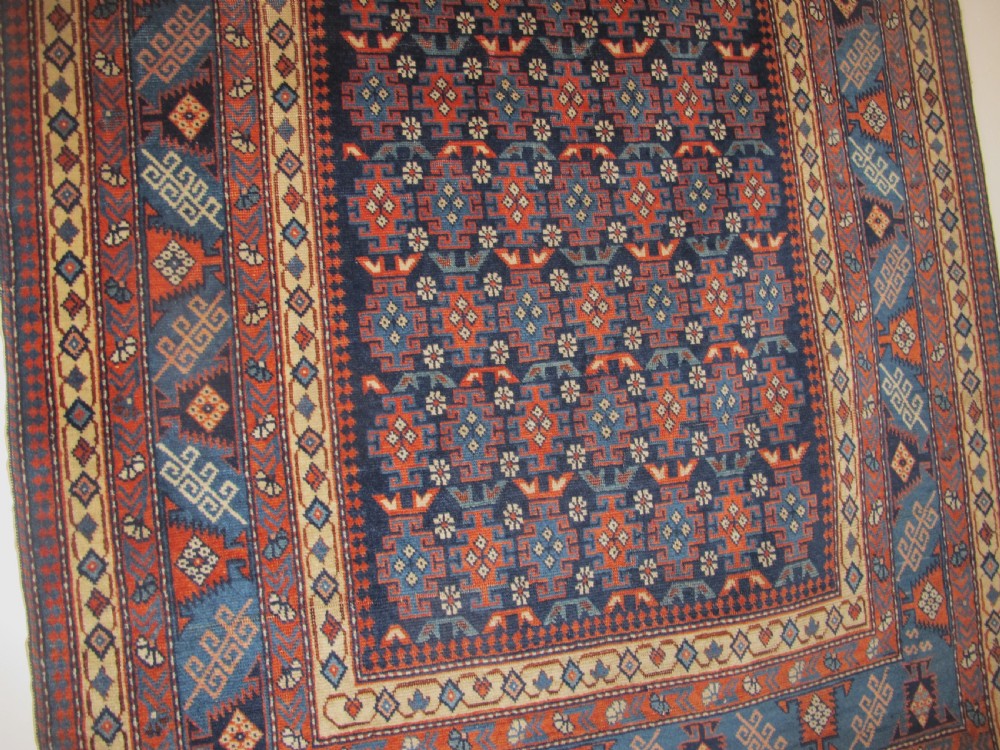 A rectangular hand-knotted wool rug featuring a dark navy central field filled with repeating geometric motifs surrounded by intricate multi-layered pattern borders in blue, terracotta, and cream. View X